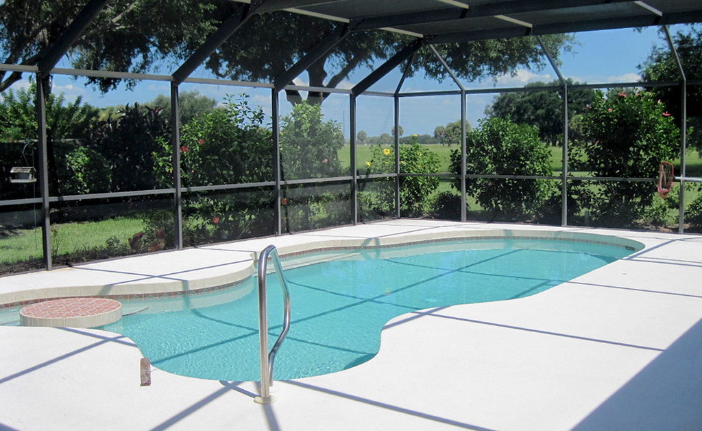 A screened pool enclosure over a swimming pool, framed in dark aluminum, with lawn and trees visible through the mesh